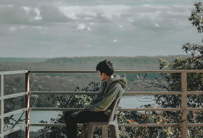 Side view of young man sitting on railing against sky