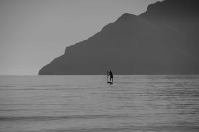 Silhouette man paddleboarding on sea against mountain during foggy weather