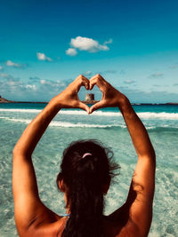 Rear view of woman making heart shape on beach against sky