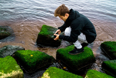 Side view of young man photographing lake while crouching on rock