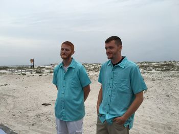 Smiling friends wearing blue shirt standing at beach against sky