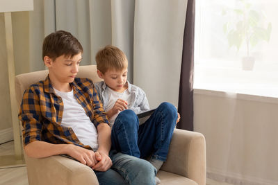 Two boys,  sit in beige armchairs, in a room, during the day, using mobile device