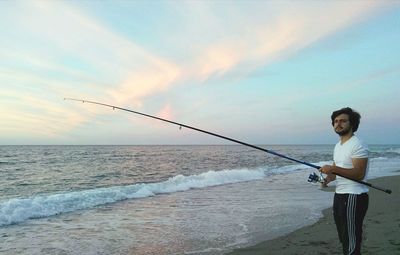 Man fishing at beach against sky during sunset