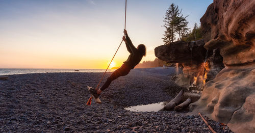 Man on rock at beach against sky during sunset