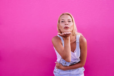 Portrait of woman blowing kiss while standing against pink wall