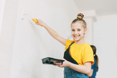 A girl in a denim overalls and a yellow t-shirt helps to paint the walls in an apartment white.