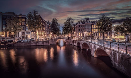 Bridge over river by buildings against sky at dusk