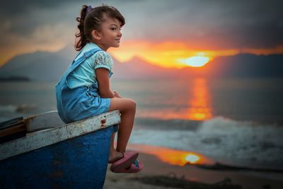 Cute girl sitting on boat at beach against sky during sunset