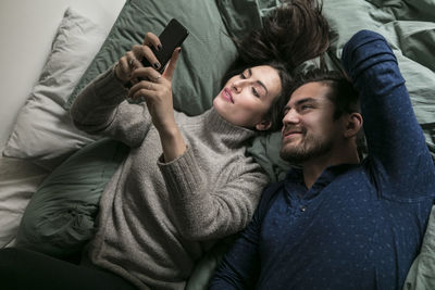 High angle view of smiling couple taking selfie through smart phone while lying on bed