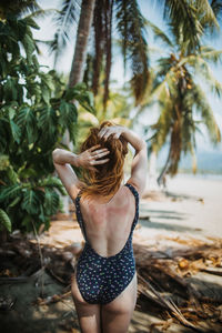 Back view of unrecognizable redhead female traveler in swimwear touching hair while standing near green palm trees on sandy beach during summer vacation in costa rica