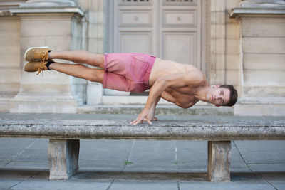 Portrait of shirtless young man balancing on bench against building
