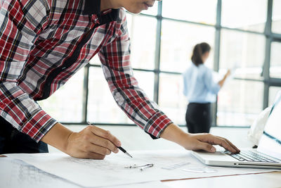 Midsection of architecture drawing blueprint while colleague working in background