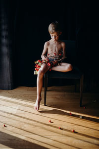 Full length of shirtless boy holding berries while sitting on chair at home