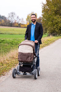 Portrait of man smiling while pushing baby stroller on road against trees