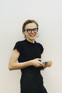 Portrait of smiling androgynous businessman leaning on white wall while holding smart phone in board room