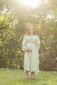 Young pregnant woman puts hands on big belly, green trees,sky on background. brown haired female