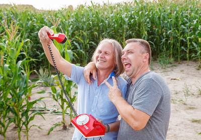 Cheerful male friends with rotary phone standing against plants in farm