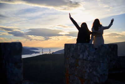 Rear view of women with arms raised standing against sky during sunset