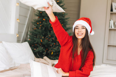 Young asian woman in cozy red sweater and santa hat having fun in room with christmas tree at home