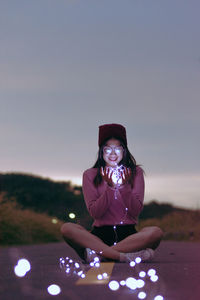 Woman holding illuminated string lights while sitting on road at dusk