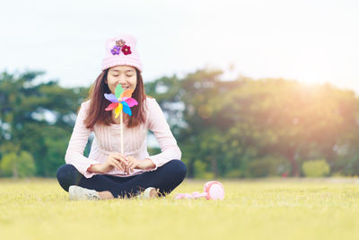 Smiling woman with closed eyes holding pinwheel while sitting on grassy field