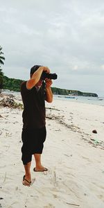 Man photographing through camera while standing on sand at beach