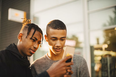 Teenage boy showing mobile phone to friend in city during sunset