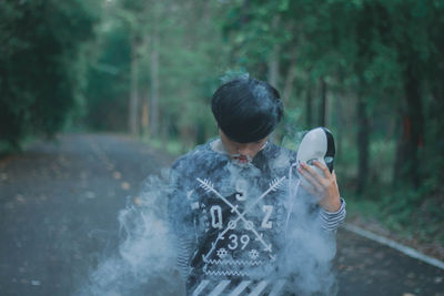 Young man holding mask with smoke while standing against trees