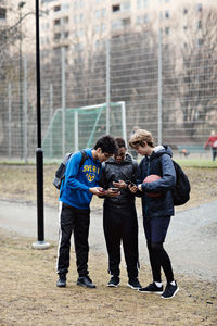 Full length of social media addicted friends wearing sports clothing while using mobile phones on playing field