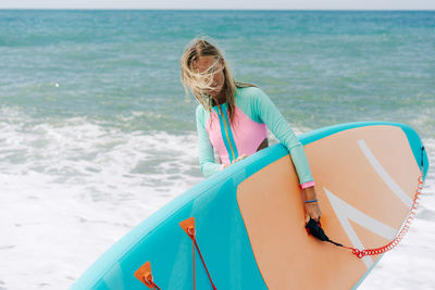 A young athletic blonde woman stands on the beach and holds a paddle board.