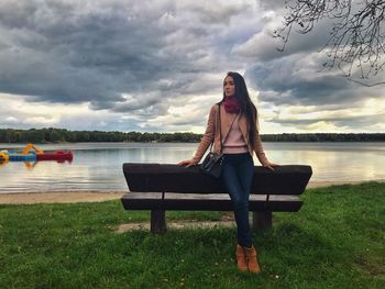 Full length of woman leaning on bench at lakeshore against sky