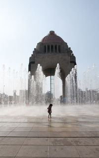 Girl standing by fountain in city against clear sky