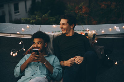 Smiling man with mobile phone sitting by friend looking away on terrace in city