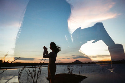 Silhouette woman photographing by sea against sky during sunset