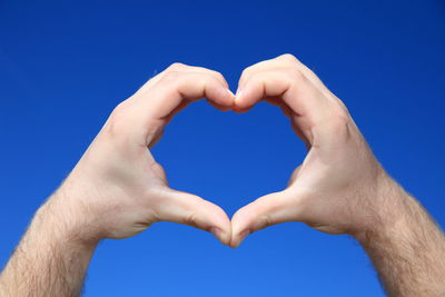 Cropped hand of man making heart shape against clear blue sky