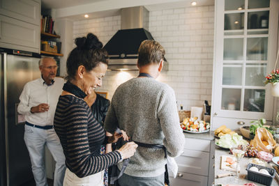 Smiling woman helping friend tying apron in kitchen at home