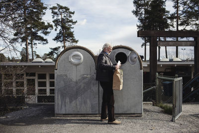 Senior man putting garbage in recycling bin against sky