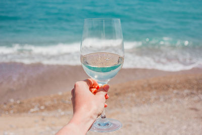 Woman hand with white wine glass on a turquoise mediterranean sea foam, waves and beach background