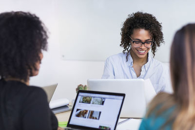 Smiling businesswomen using laptops while sitting with colleagues in meeting at board room