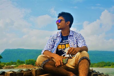Young man wearing sunglasses while sitting on mountain against sky