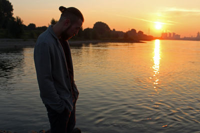 Side view of young man standing by lake against sky during sunset