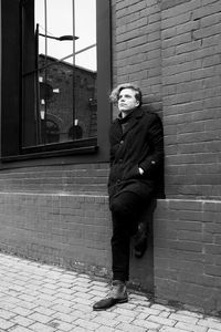 Full length of thoughtful young man looking away while leaning against brick wall