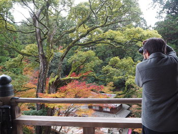 Rear view of man photographing trees while standing by railing in balcony