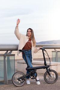 Vertical view of young woman waving at someone while holding an electric bicycle 