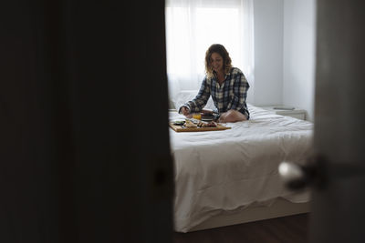 View thru a door ajar of woman having breakfast on bed.