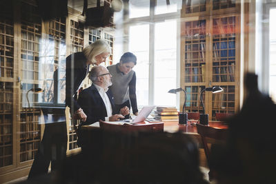 Senior lawyer discussing with female coworkers at table seen through glass in library