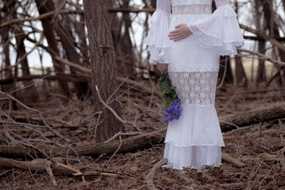 Low section of bride holding purple flowers by bare trees in forest