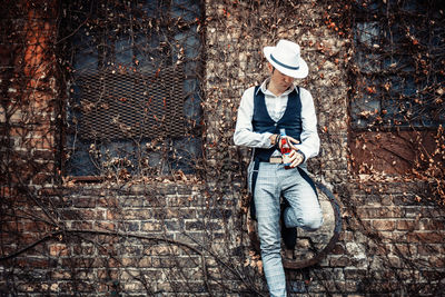 Portrait of retro-styled man holding beer bottle while adjusting sleeve and leaning on a wall.