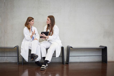 Full length of confident female doctors discussing over digital tablet while sitting against wall at hospital