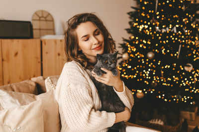 Smiling happy girl hugging her pet cat while sitting by a decorated christmas tree in a cozy home